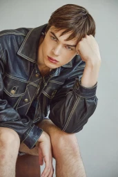 A young man seated in a black leather jacket with contrast stitching rests his head on his hand against a neutral studio backdrop.