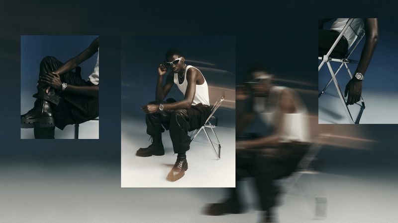 A man wearing a white tank top and dark trousers sits on a folding chair, sporting sunglasses and a wristwatch in a studio setting.