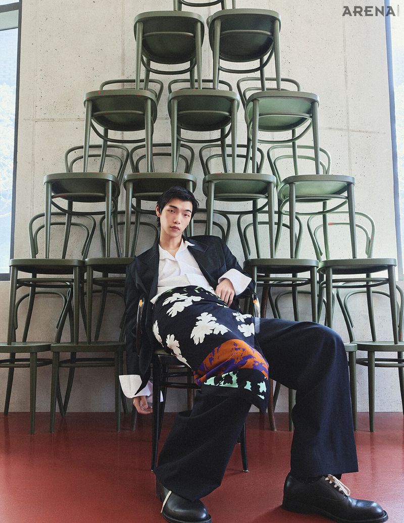 Model Suyong Jung seated in dark tailoring and a floral wrap before a pyramid stack of green chairs.