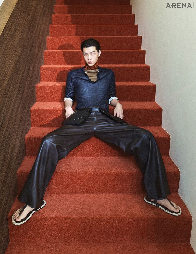 East Asian male model wearing a dark blue polo and wide-leg leather trousers sitting on red carpeted stairs, looking at camera.