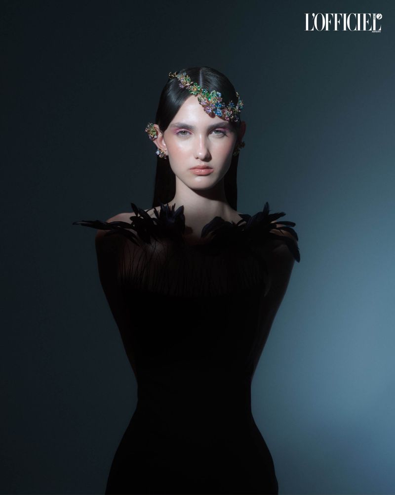 A fashion portrait of a young woman with a jeweled headpiece and black feathered dress against a dark blue gradient background.