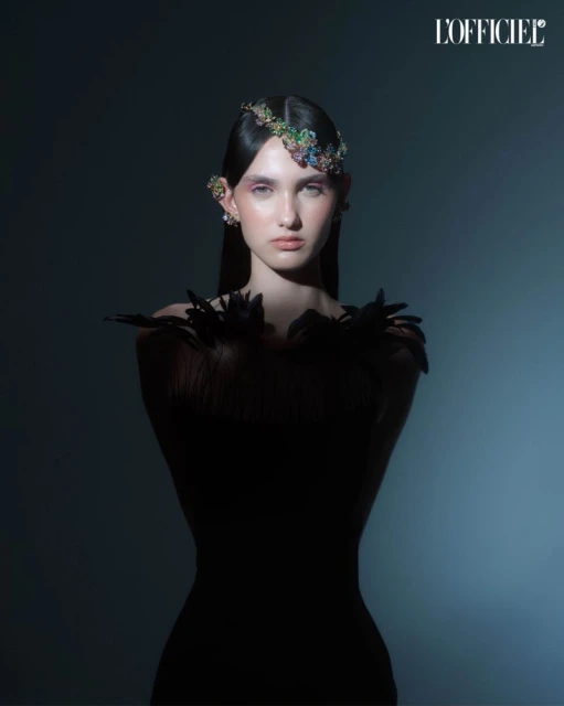 A fashion portrait of a young woman with a jeweled headpiece and black feathered dress against a dark blue gradient background.