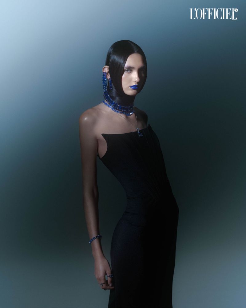 A striking fashion portrait features a model wearing a form-fitting black dress and bold blue jewelry and makeup against a blue gradient.