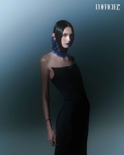 A striking fashion portrait features a model wearing a form-fitting black dress and bold blue jewelry and makeup against a blue gradient.