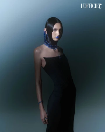 A striking fashion portrait features a model wearing a form-fitting black dress and bold blue jewelry and makeup against a blue gradient.