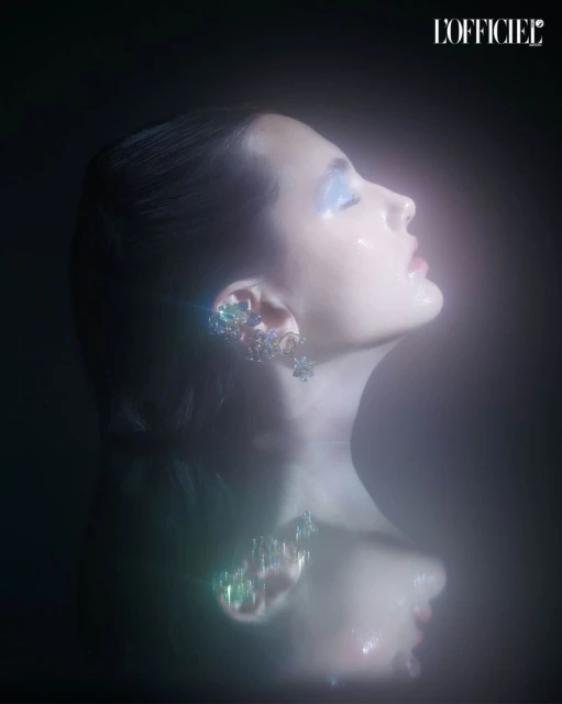 Profile shot of a woman with blue eyeshadow and jeweled ear cuffs, looking upwards against a dark background with a reflection.