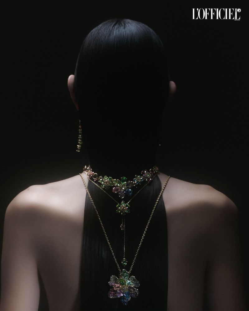 Back view of a woman with a dark bob haircut wearing an ornate necklace with crystal flowers and gold chain in a dark studio.