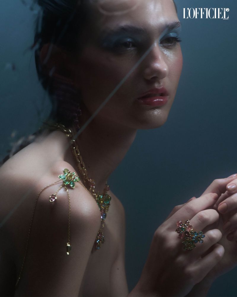 Model with blue eye makeup, gold necklace, and ring poses in a close-up studio shot with a dark blue background and fashion magazine logo.