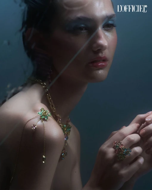 Model with blue eye makeup, gold necklace, and ring poses in a close-up studio shot with a dark blue background and fashion magazine logo.