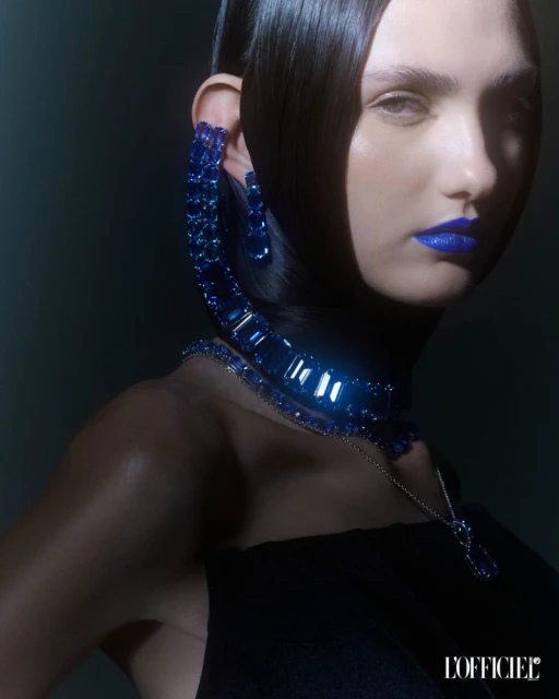 Close-up beauty shot of a fair-skinned woman with sleek black hair, dramatic blue lipstick, and matching blue gemstone jewelry.