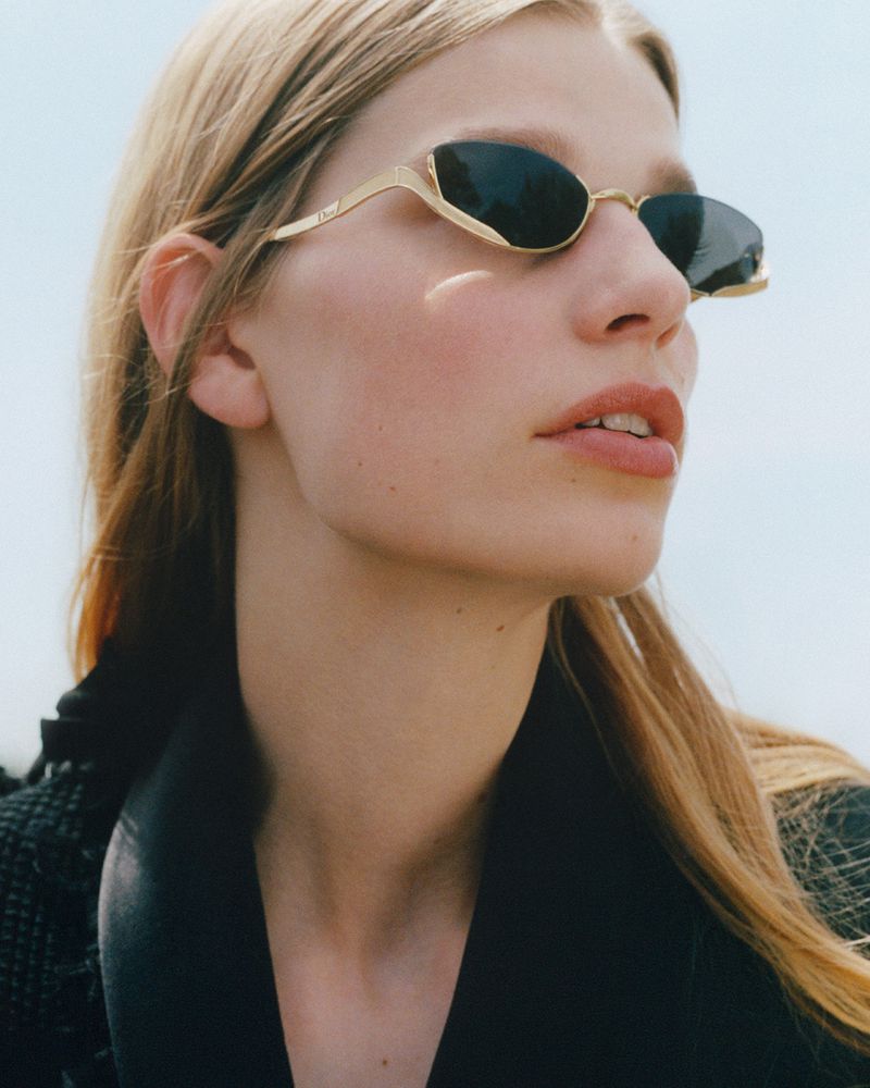 A close-up portrait of a woman with blonde hair wearing dark Dior sunglasses with gold frames against a bright, hazy sky.
