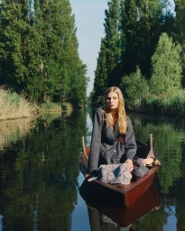 A blonde woman in a grey double-breasted coat sits in a wooden rowing boat on a tree-lined canal with a patterned handbag.