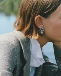 Close-up profile of a woman with a bob haircut wearing a grey tweed blazer, white shirt, and a crystal bow earring by water.