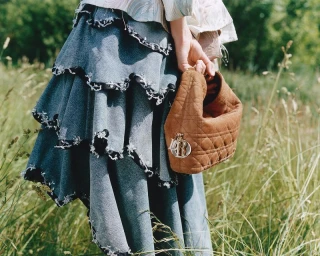 Close-up of a tiered blue denim ruffled skirt with raw frayed edges and a brown quilted suede handbag held in a grassy field.