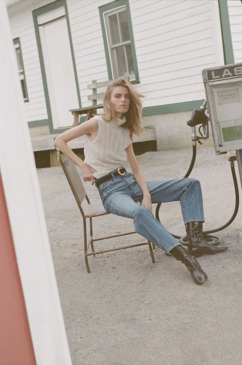 A fair-skinned blonde woman in a sleeveless cream knit top and blue jeans sits on a chair near an old gas pump.