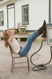 Model Marie-Violaine Chenal lounging in a metal chair with tall brown leather boots propped on a vintage gas pump.