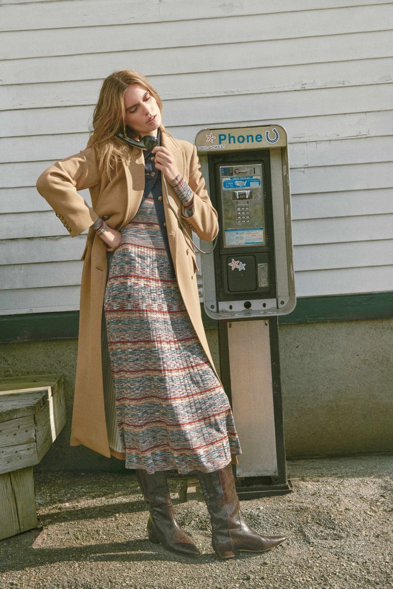 Model Marie-Violaine Chenal stands by an outdoor payphone wearing a long camel coat, striped knit dress, and brown knee-high boots.