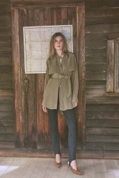 Marie-Violaine Chenal stands before a rustic wooden door wearing an olive belted tunic, black trousers, and pointed heels.