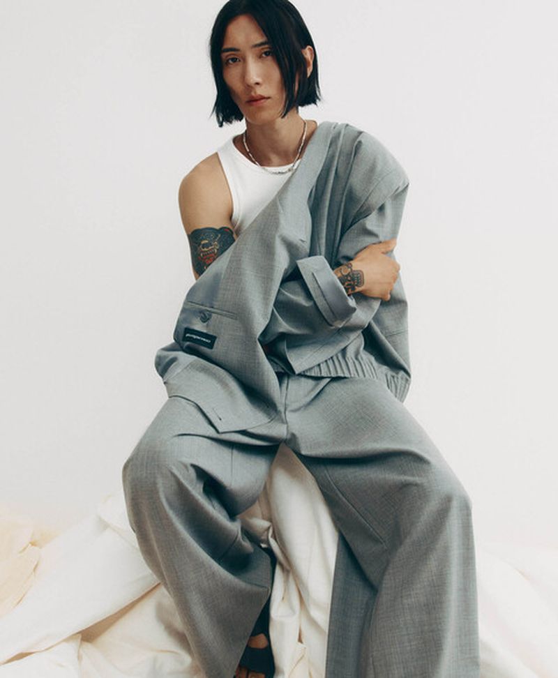 Model Hyuk Kim seated in a draped oversized grey suit and white tank top against a plain studio background for Adolfo Dominguez.