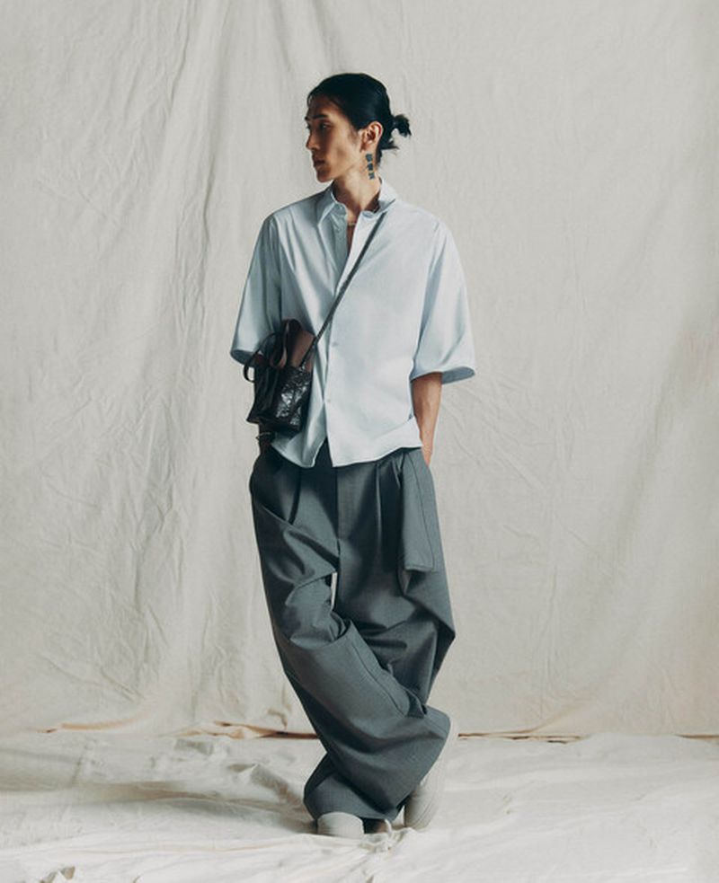 Model Hyuk Kim posing in a light blue short-sleeved shirt and oversized grey trousers against a textured white fabric backdrop.