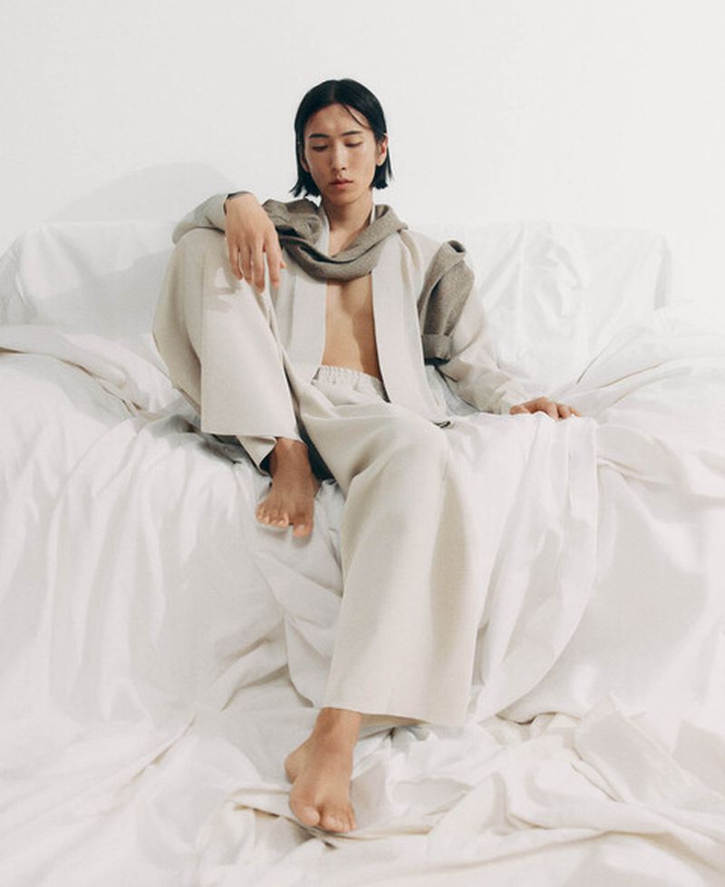 Model Hyuk Kim seated barefoot in wide-leg cream trousers and an open robe draped in white fabric for Adolfo Dominguez.