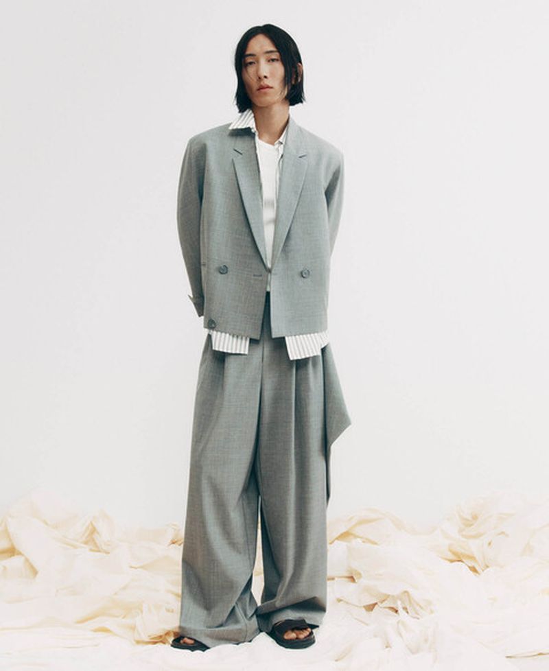 Model Hyuk Kim wearing an oversized grey suit and black sandals standing on draped fabric against a white studio background.