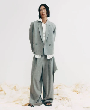 Model Hyuk Kim wearing an oversized grey suit and black sandals standing on draped fabric against a white studio background.