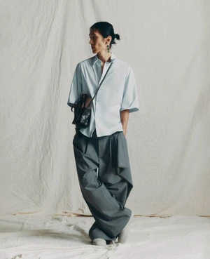 Model Hyuk Kim posing in a light blue short-sleeved shirt and oversized grey trousers against a textured white fabric backdrop.