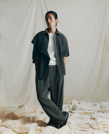 Model Hyuk Kim wearing a dark pleated shirt and matching trousers against a draped fabric backdrop.