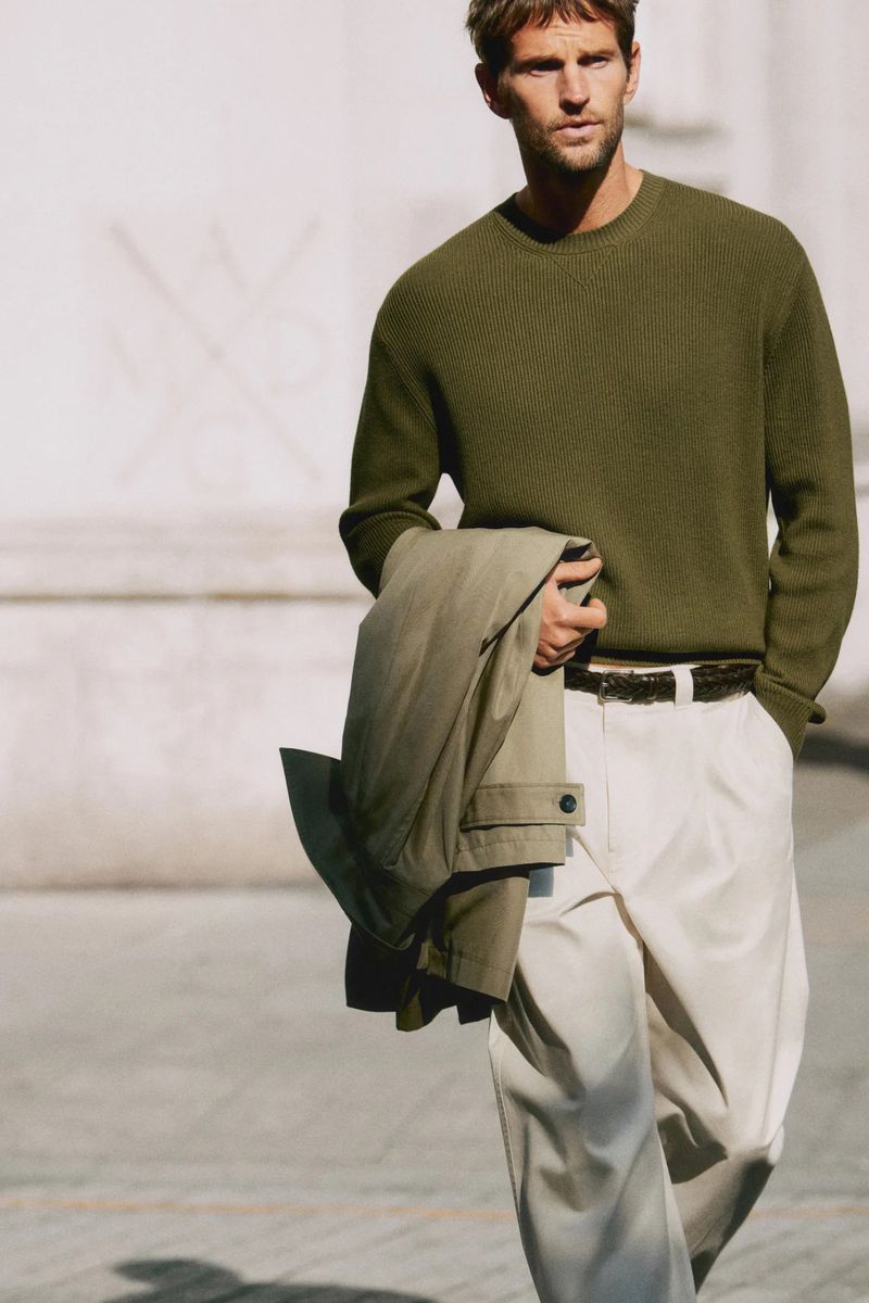 Man wearing a long-sleeved olive green sweater and cream pleated trousers carries a folded beige coat outdoors in daylight.