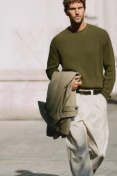 Man walking in an olive green knit sweater and pleated off-white trousers carrying a folded trench coat on his arm outdoors.