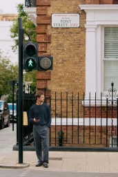 Man in a dark textured sweater and grey trousers standing beside a pedestrian signal on Pont Street.