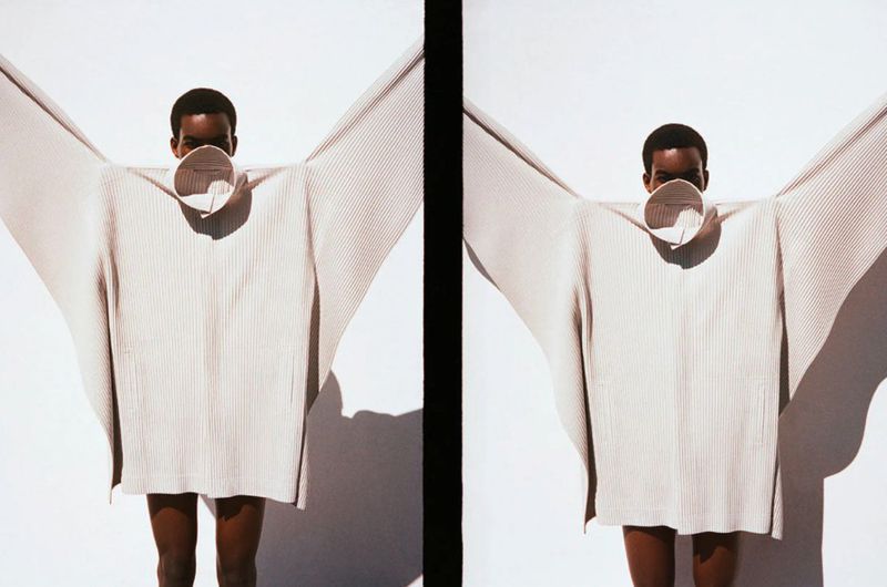 A person wearing a beige pleated Issey Miyake garment that extends outwards like wings against a stark white background.