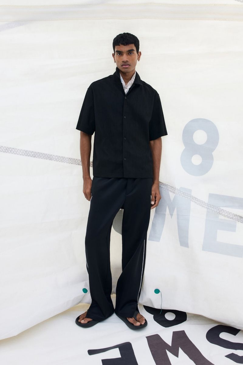 A young South Asian man stands before a white sail, wearing a black short-sleeved button-up shirt, matching black trousers, and sandals.