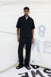 Model Deepak Gupta poses in a black short-sleeved shirt, wide-leg trousers, and sandals in front of a white sail backdrop.