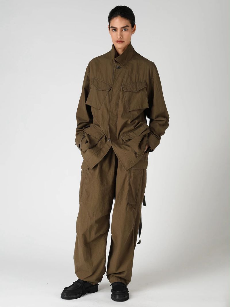A person with short hair wears an olive-brown utility jacket and matching wide-leg cargo trousers against a plain light grey background.