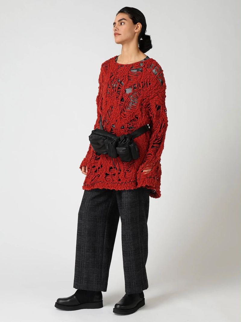 Model wearing a distressed red knit sweater, charcoal wide-leg trousers, and a black belt bag against a white backdrop.