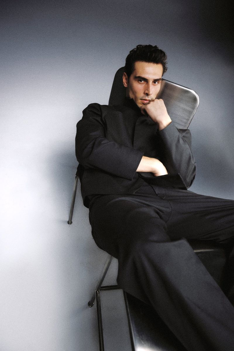 A man seated in a metal chair wearing a tailored black suit against a softly lit grey studio backdrop.
