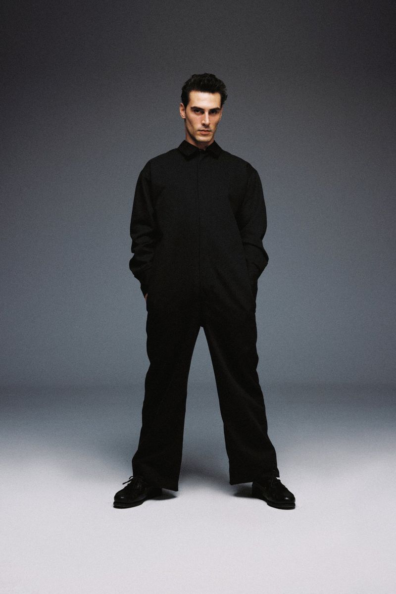 Model Eren Semerci standing in a full-length black collared jumpsuit and black shoes against a grey studio backdrop.