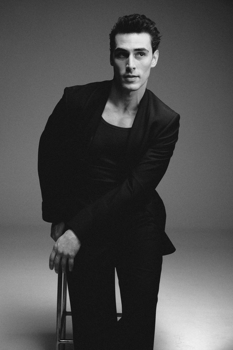 A young man with dark, styled hair wears a black blazer and tank top while posing on a stool in a studio against a plain background.