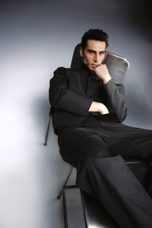 A man seated in a metal chair wearing a tailored black suit against a softly lit grey studio backdrop.