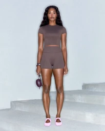 A Black woman in a brown short-sleeve crop top and matching biker shorts stands on concrete stairs wearing pink flats.