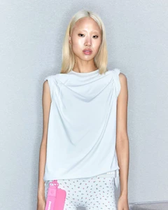 East Asian woman with bleached blonde hair posing in a sleeveless pale grey draped top and printed leggings against a studio wall.
