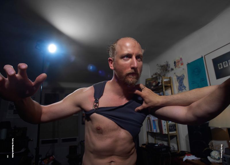 Man in black tank top strikes a dramatic pose indoors with extended arms. Another person's arm is on his shoulder, indoor setting.