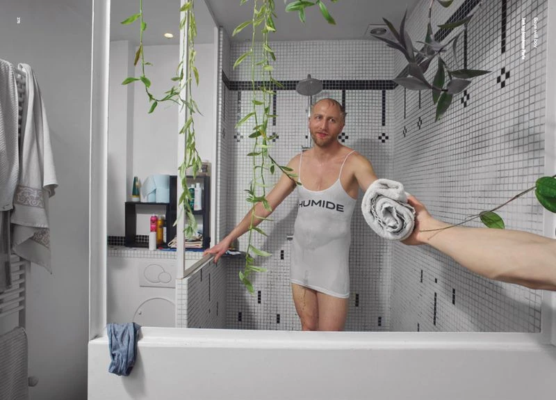 Adult male in bathroom wearing a wet tank top holding a towel. Modern, tiled bathroom with plants and laundry appliances.