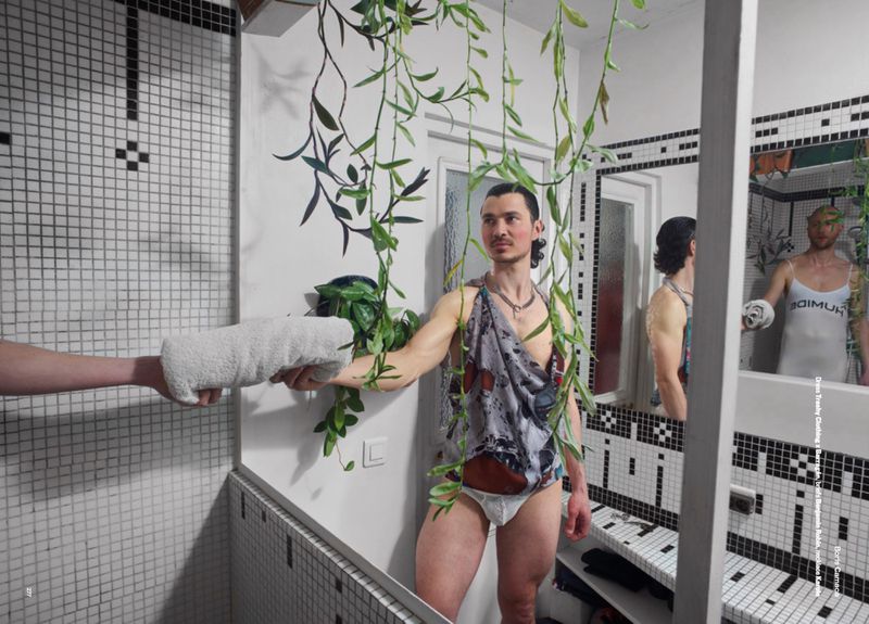 Man posing in a bathroom mirror in briefs and a toga-like garment, a hand offers a towel; verdant vines and black-and-white tile setting.