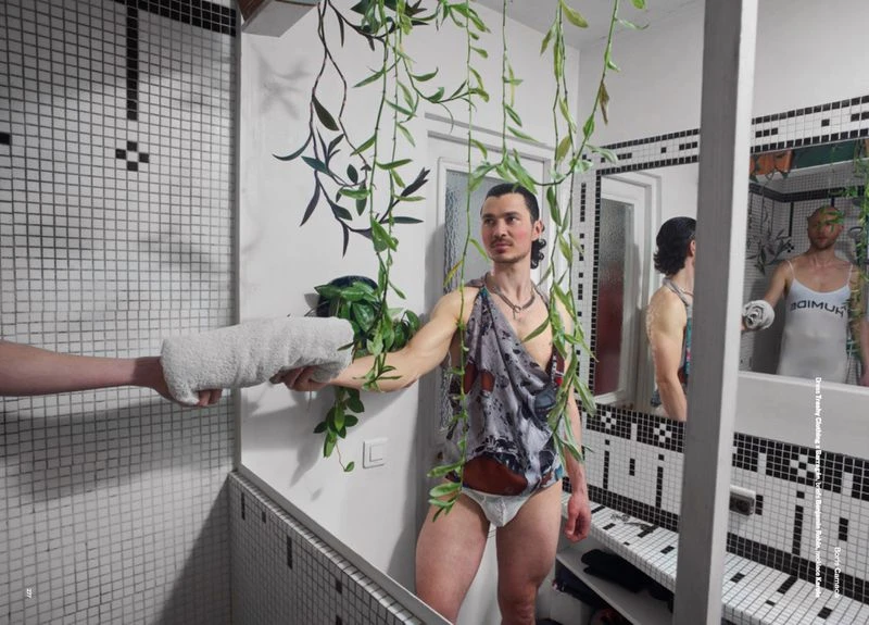 Man posing in a bathroom mirror in briefs and a toga-like garment, a hand offers a towel; verdant vines and black-and-white tile setting.