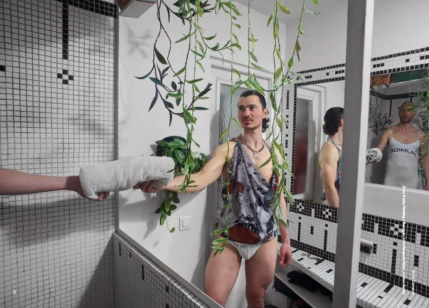 Man posing in a bathroom mirror in briefs and a toga-like garment, a hand offers a towel; verdant vines and black-and-white tile setting.