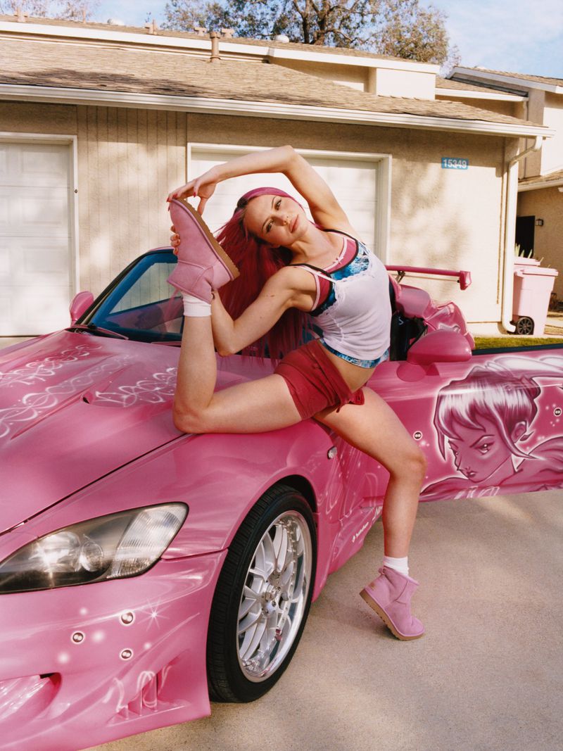 Woman with pink hair posing in a split on the hood of a custom pink car, wearing a white tank top, red shorts, and pink Ugg boots.