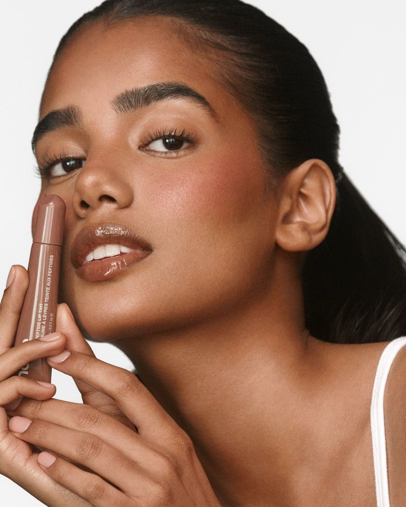 Close-up beauty portrait of a woman holding a brown lip tint tube next to her glossy lips against a warm studio backdrop.